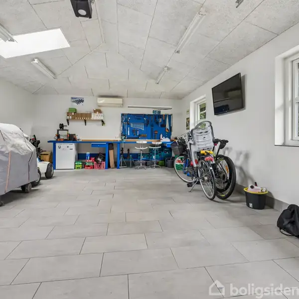En garage med en motorcykel dækket af en presenning, cykler parkeret ved siden af, og en arbejdsbænk fyldt med værktøj. Et vindue til højre giver lys til rummet.