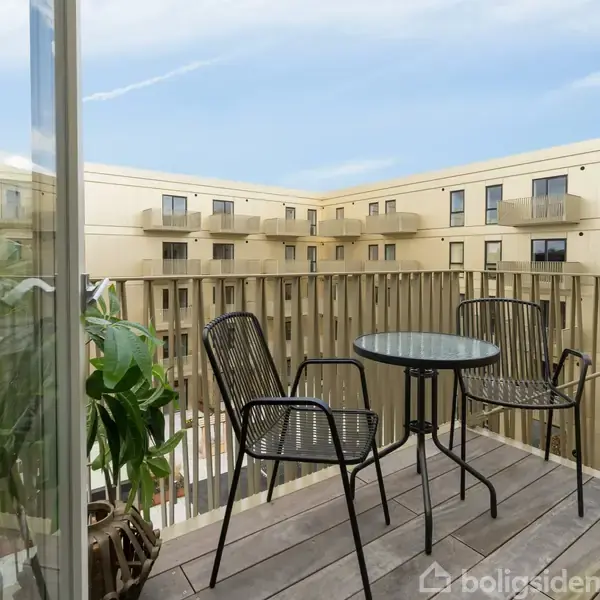 En balkon med to sorte stole og et rundt bord står i et boligkompleks med beige bygninger omkring. En stor plante står ved balkonkanten.