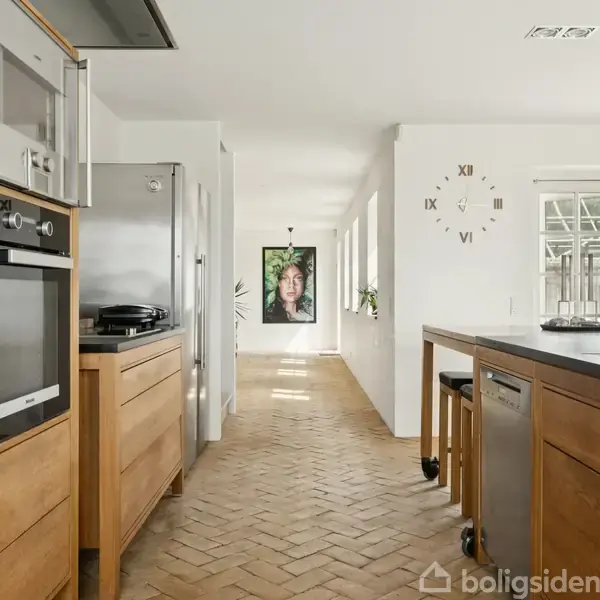 Køkken med træskabe og moderne apparater langs væggen. En stor ur med romertal hænger på væggen. Gennem vinduerne indledes udsigt til en have.