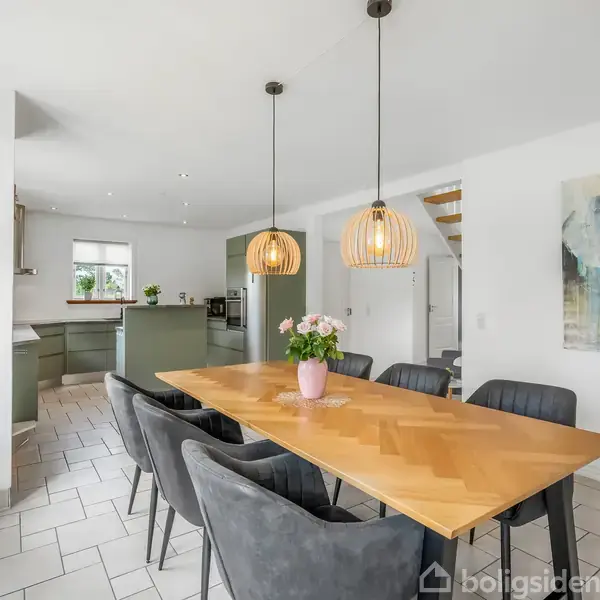 Et spisebord er omgivet af seks mørke stole og har en lyserød vase med blomster. Det står i et lyst køkken-alrum med lysekronelamper og moderne køkken i baggrunden.