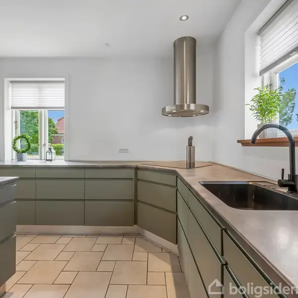 Et moderne køkken med grønne skabe omgiver en sort vask og køkkenhane. Et stort vindue med planter står i baggrunden, og der er en emhætte over komfuret. Skakternet gulv.