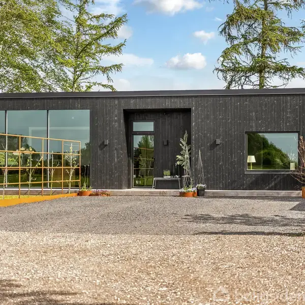 En moderne, sort træbeklædt bygning med store vinduer står stille i en have med grusbelagt indkørsel og grønne træer i baggrunden.