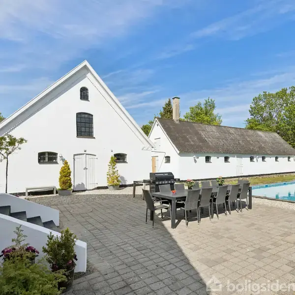 Et langbord med stole står på en flisebelagt terrasse ved siden af en swimmingpool, omgivet af grønne træer og to hvide bygninger med skrå tage under en blå himmel.