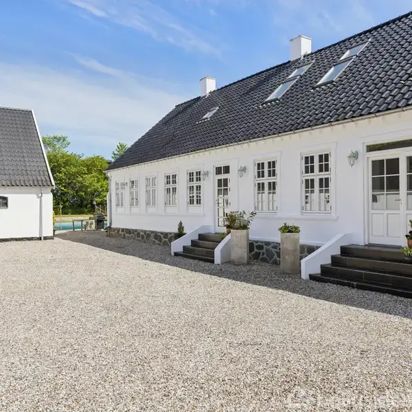 En hvid, traditionel bygning med sorte tegltage står ved siden af en større gårdsplads af perlesten. Bygningen har flere symmetriske vinduer og en havedør med trapper.