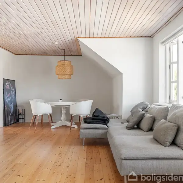 Grå sofa med puder står ved vindue. Rundt bord med hvide stole står under lampeskærm. Trægulv og træloft fremhæver den lyse stue.