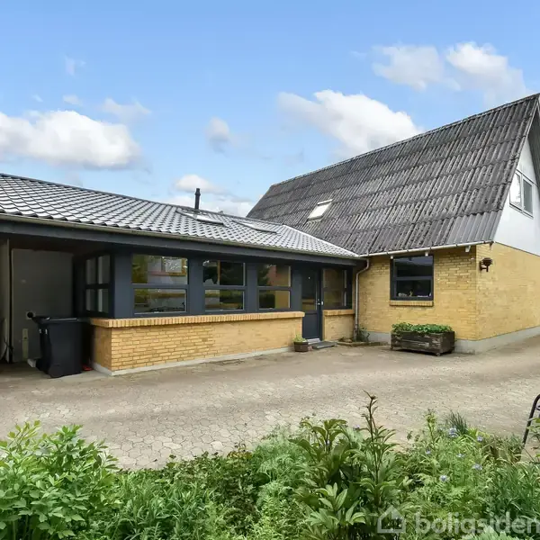 Hus med gule mursten og grå tagsten står på en gårdsplads. Der er en grøn have i forgrunden og blå himmel med få skyer i baggrunden.