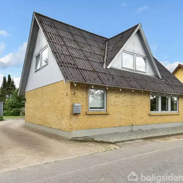 Gult murstenshus med skråt tag står ved en vej. Postkasse ved hoveddøren, omgivet af grønne hække og træer i baggrunden. Skyet blå himmel i baggrunden.