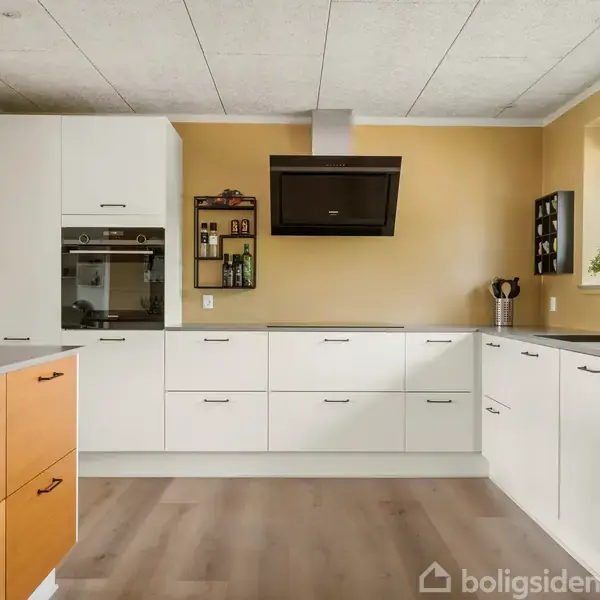 Hvidt, moderne køkken med integrerede skabe og ovn. Et ventilationssystem hænger centralt. Køkkenkrukker og planter står ved et vindue på højre side. Trægulv og varm farvetone.