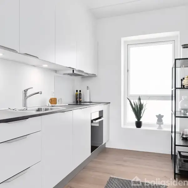 Hvidt køkken med indbyggede skabe og sort bordplade. Vask og ovn er indlejret. Der er en reol ved siden af et vindue med en plante. Gulvet er træ.