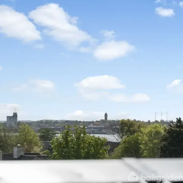 Et panoramavindue viser en byudsigt med grønne træer i forgrunden og spredte bygninger mod en blå himmel med skyer i baggrunden.