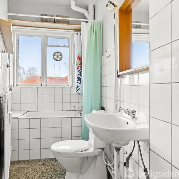 Badeværelse med flisebelagte vægge og gulv, toilet og håndvask placeret ved siden af et badekar med bruser. Et grønt bruseforhæng hænger ved vinduet, der lukker lys ind.