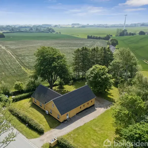 Gult hus med sort tag står blandt træer, omgivet af grønne marker og en grusvej i landlig idyl.