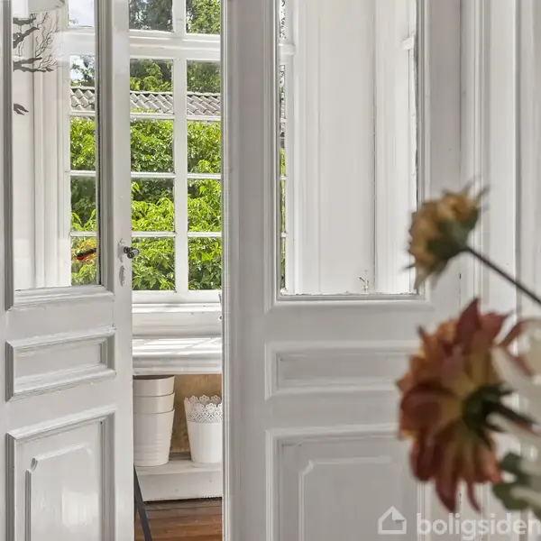 Dobbeltdøre står åbne mod en lysfyldt veranda med store vinduer, omgivet af grønne buske. I forgrunden ses blomster i vaser.
