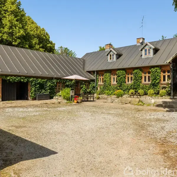 Et landsted med grusgårdsplads er omgivet af grønne træer. Huset har et mørkt skifertag og flere skrå kvistvinduer, mens garagebygningen flankerer gårdspladsen.