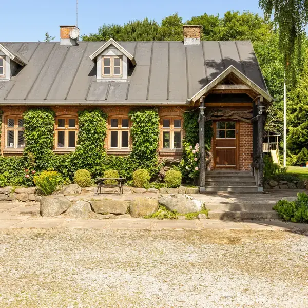 Et landsted med klatreplanter dækker facaden, og en veranda med trapper fører til hoveddøren. Omkring er der en gårdsplads med blomsterpotter og en græsplæne med træer.