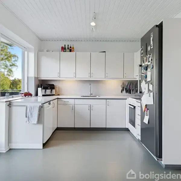 Køkken med hvide skabslåger, hvor et køleskab er fyldt med papirnoter. Udenfor ses grønt løv gennem en glasdør, som står ved siden af en grill.