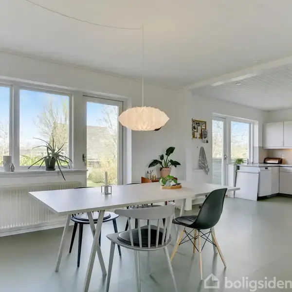 Et lyst køkkenalrum med et hvidt spisebord og sorte stole. En unik lampe hænger over bordet. Store vinduer og planter skaber en lys og indbydende atmosfære.