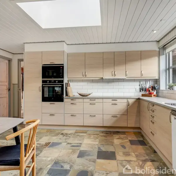 Køkken med moderne træskabe og hvide fliser som backsplash. Der er en opvaskemaskine, indbygget ovn og store vinduer, der slipper lys ind. Rundt hjørne står et spisebord med stole.