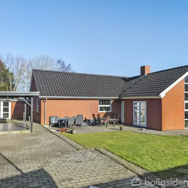 Et rødt murstenshus med store vinduer. Huset har en terrasse med havemøbler, carport til venstre, og græsplæne foran. Træer og en blå himmel i baggrunden.