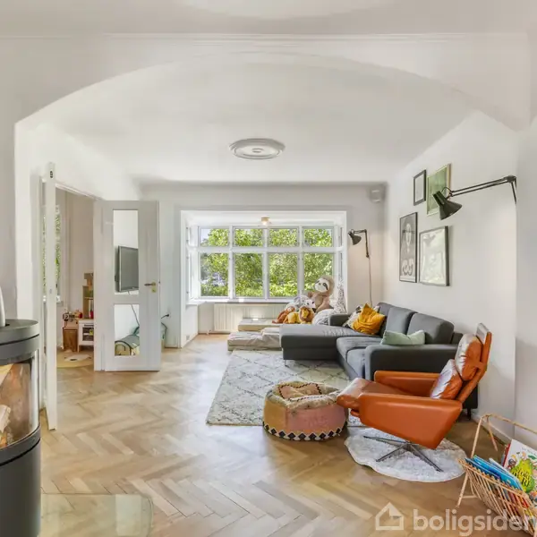 Stue med grå sofa og orange lænestol placeret på trægulv, pejs til venstre. Store vinduer i baggrunden giver udsigt til grøn beplantning udenfor. Hyggelig og lys atmosfære.