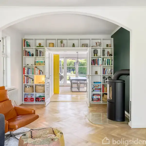 En stue med en brun lænestol og en reolvæg fyldt med bøger; en brændeovn står ved siden af. Åbne døre fører til et rum med et spisebord og glasdøre til haven.