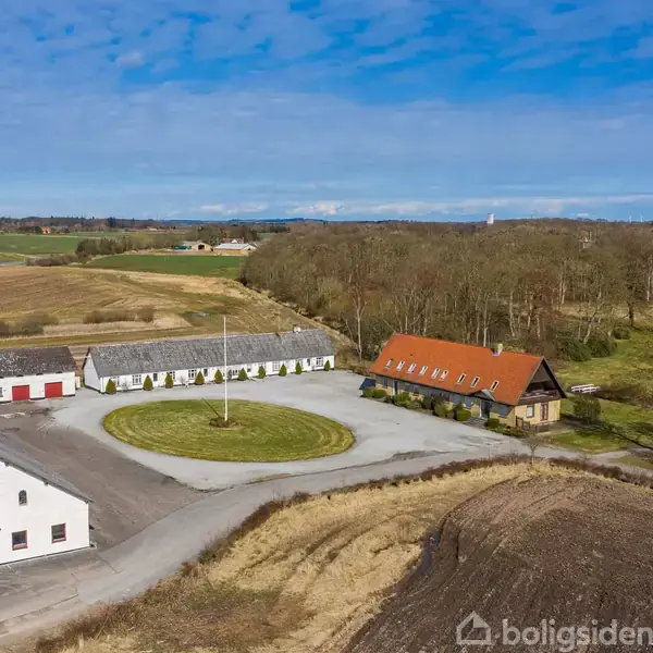 Gårdsplads omgivet af flere bygninger, herunder en hvid stald og en rødtag villa. Rundt græsareal omkranser flagstang. Omringet af marker og skovklædte områder under klar blå himmel.