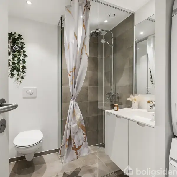 Badeværelse med brusekabine, badeforhæng og vægmonteret toilet, omgivet af lyse fliser. Til højre ses en vaskemaskine og tørretumbler samt en håndvask med spejl.