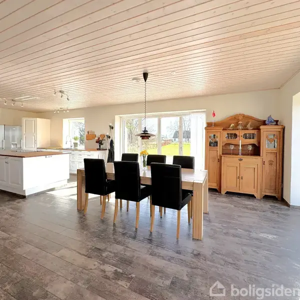 Spisebord med seks sorte stole står i et lyst rum med træloft. Til højre ses et vitrineskab, og i baggrunden er et køkken med ø. Store vinduer giver udsigt til en have.