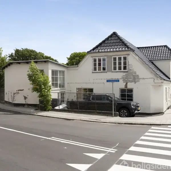En hvid bygning med skråt tag står ved et vejkryds. En sort bil er parkeret foran. I baggrunden er træer. Skiltet viser "Silkeborgvej".
