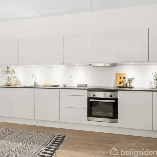Et moderne køkken med hvide skabe og en sort bordplade. Oven på bordpladen står en plante, skærebræt og køkkenredskaber. Et geometrisk mønstret tæppe ligger på det lyse trægulv.