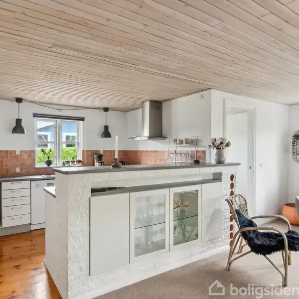 Køkkenø med barstole står i et moderne køkken med hvidmalede lofter og træparket. Køleskab og indbygningsskabe ses til venstre, med en sofa og vinduer i baggrunden.