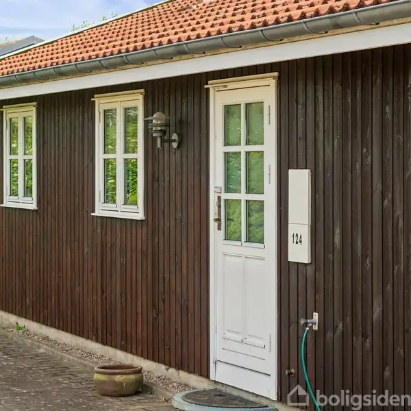 En brun bygning med hvid dør og to vinduer vises. Døren har nummeret 124. Bygningen er omkranset af grønne buske, og flisebelægningen er udenfor.