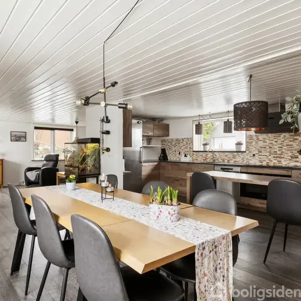 Spisebord omgivet af sorte stole, dekoreret med dug og planter, står i et lyst, åbent køkken-alrum med træmøbler. I baggrunden ses en sofa og reolsystem.