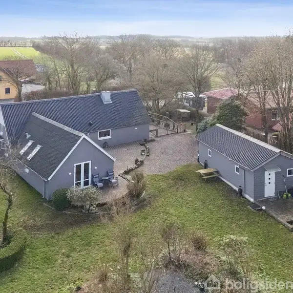 Gråt hus med to etager og sort tag står i en have med træer og græs, ved siden af et mindre gråt udhus. Omgivelserne inkluderer flere bygninger og åbne marker.