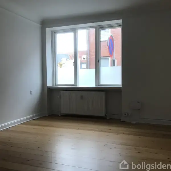 Et tomt værelse med trægulv har stort vindue med udsigt til en murstensbygning. En radiator er under vinduet, og væggene er hvide.