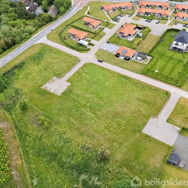 Grønt, åbent græsareal ligger stille tæt på et boligområde med rødtagehuse og veje, omgivet af grønne marker og et lille industriområde.