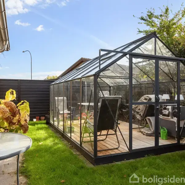 Et drivhus med glasvægge står på en træterrasse, indeholdende havemøbler. Det er placeret i en grøn have ved siden af et murstenshus, omgivet af buske og blomster.
