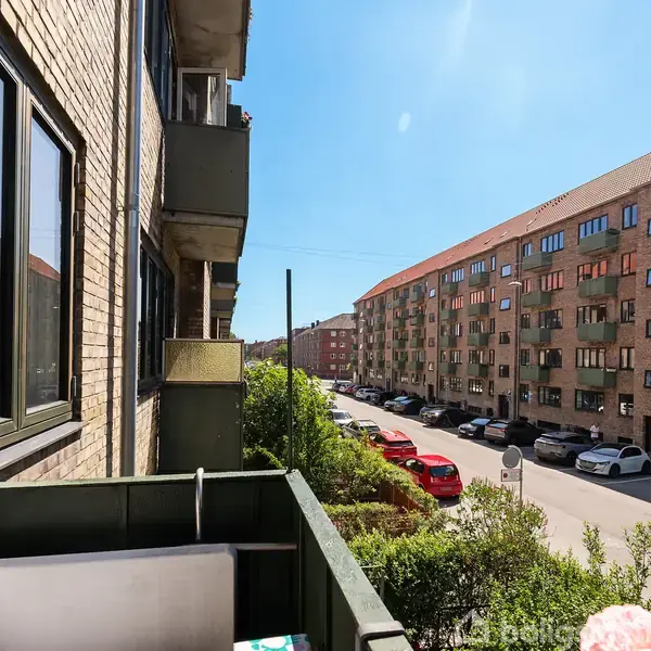 En mindre altan med blomstrede puder og haveudsigt. Til højre ses parkerede biler langs en rolig gade med række af murstensbygninger under en klar blå himmel.