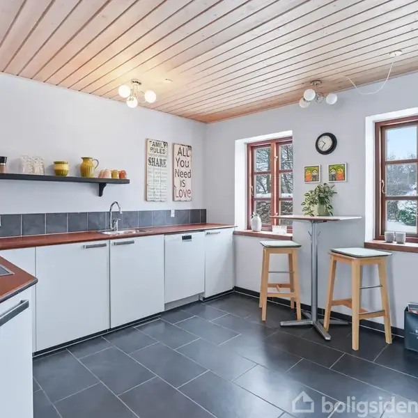 Et køkken med hvide skabe og rødbrune bordplader, hvor væggen har hylder med krukker. To vinduer med barbord og skamler. Tekst: "ALL YOU NEED IS LOVE."