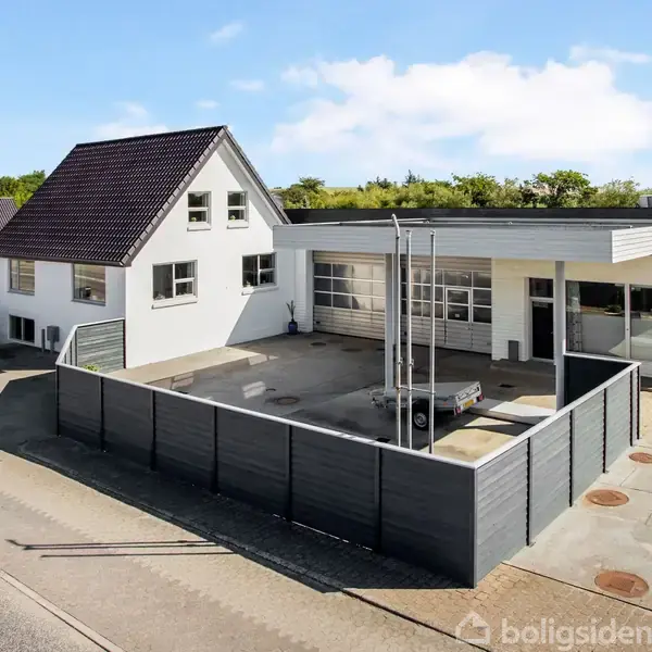 Hvid to-etagers bygning med sort tag står stille ved siden af en flad bygning med store vinduer og garageport. Omgivet af en lav, sort mur med adgang til vejen.