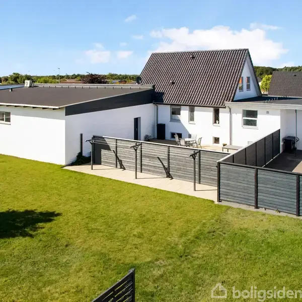 Hus med hvid facade og mørkt tag står på en grøn græsplæne. Ved siden af er en terrasse med hegn og borde. Blå himmel og få skyer i baggrunden.