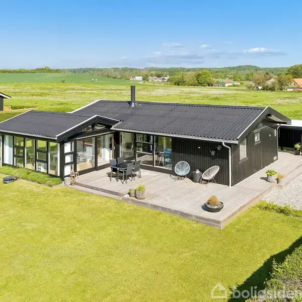 Sort enfamiliehus står stille på en åben græsplæne med en stor terrasse, omgivet af marker og flere huse i det fjerne under en blå himmel.