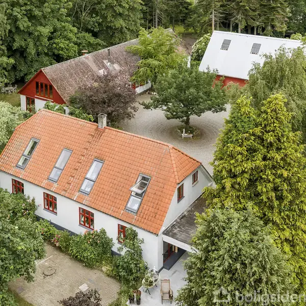 Hus med rødt tegltag og hvide mure, omgivet af træer og en gårdsplads. To andre større bygninger og biler ses i baggrunden. Skovområdet omgiver ejendommen tæt.