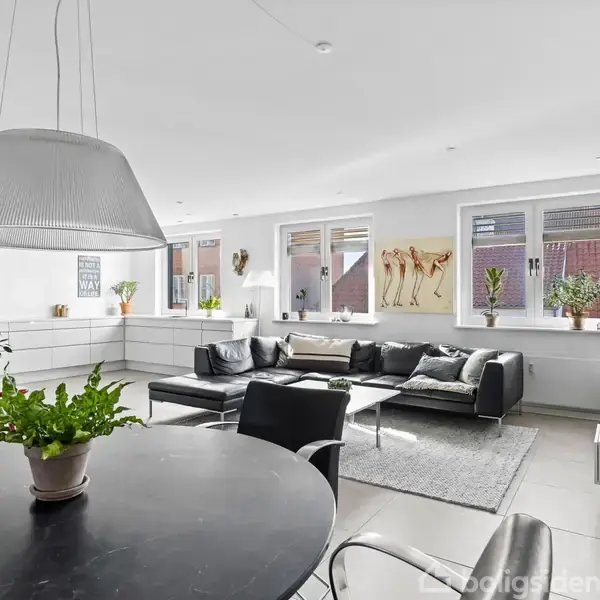 En moderne og lys stue med en sort lædersofa og et gråt tæppe. Spisebordet i forgrunden har en grøn plante, mens store vinduer slipper masser af lys ind.