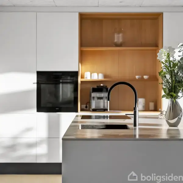 En køkkenø med en sort vandhane står i forgrunden. I baggrunden er der indbyggede skabe og en hylde med kaffemaskine og kopper, oplyst af naturligt lys.