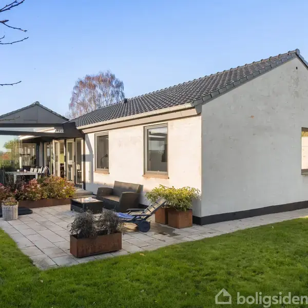 Et hvidt hus med sort tag ligger i en grøn have. Foran huset er en terrasse med møbler og planter, omgivet af græsplæne og træer i baggrunden.