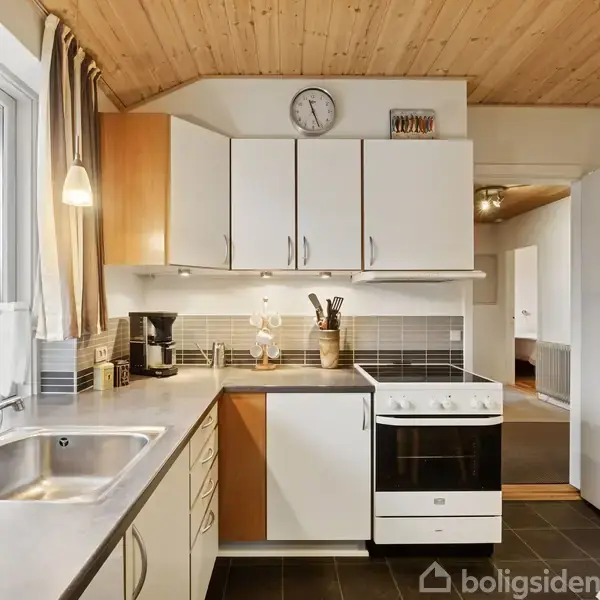 Et lyst køkken med hvide skabe hænger over en komfur og opvaskemaskine. Vask og kaffemaskine står ved et vindue. Træloft og flisegulv fuldender rummet.