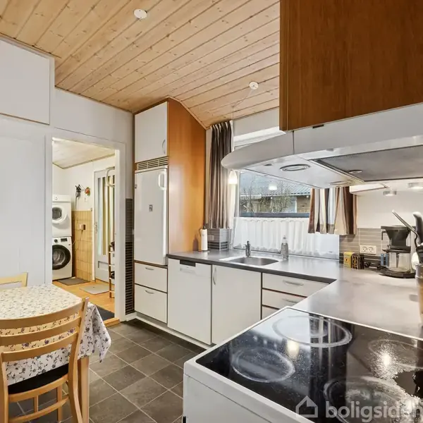 Et køkken med hvide skabe og en sort komfur, omgivet af trævægge og -loft. Spisebord med blomstret dug og fire stole står ved siden af. Vaskerum ses i baggrunden.