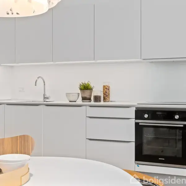 Moderne køkken med hvide skabe og indbygget ovn. Bordplade med håndvask, dekoreret med planter og krukker. Et rundt spisebord med træstole står i forgrunden og en lampe hænger over bordet.