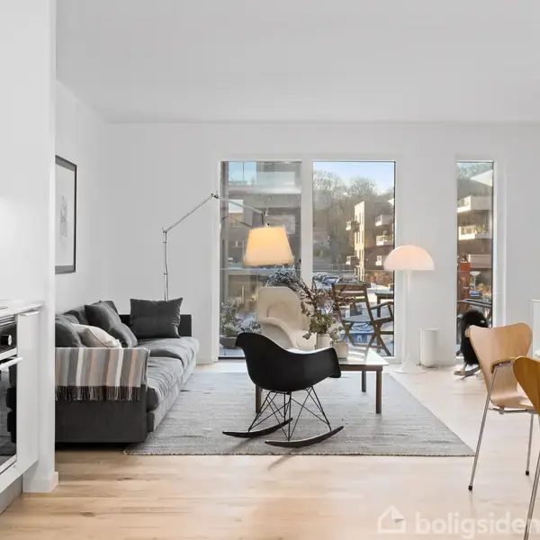 Stue med moderne indretning. En sofa og gyngestol står på et tæppe foran store vinduer. Et spisebord med stole befinder sig til højre. Køkken ses til venstre.